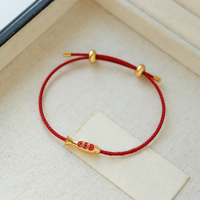 Dainty red string bracelet with brass koi charm on beige fabric surface