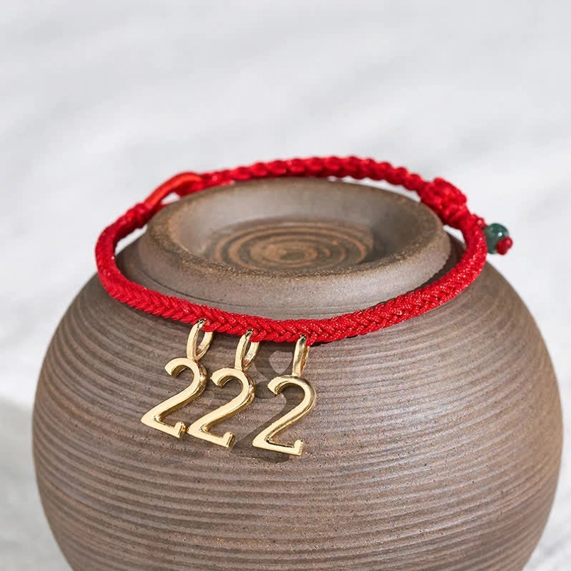 Angel number red string bracelet detail view showing woven red rope, gold number pendant, and green jade accent bead on ceramic backdrop