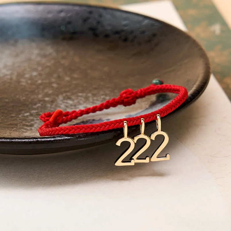 Angel number protection bracelet side view, red woven cord with gold charm pendant and green jade bead on dark ceramic plate