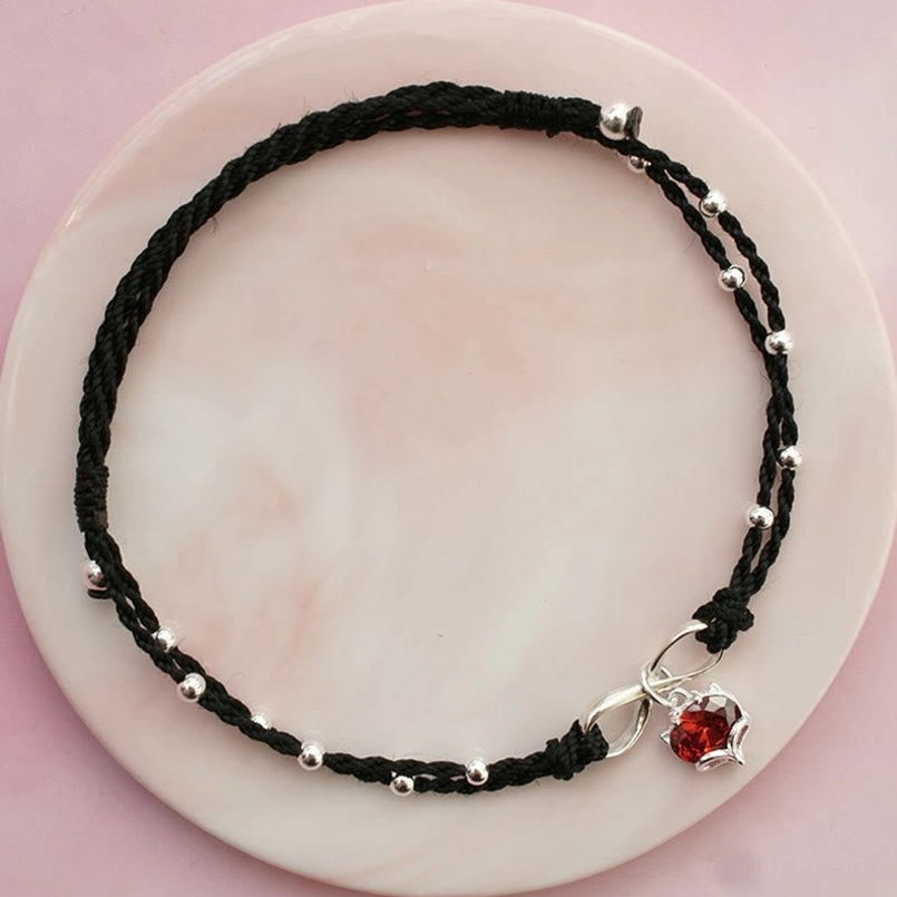 Black braided string ankle bracelet with heart charm and infinity symbol on pink ceramic plate, black variant