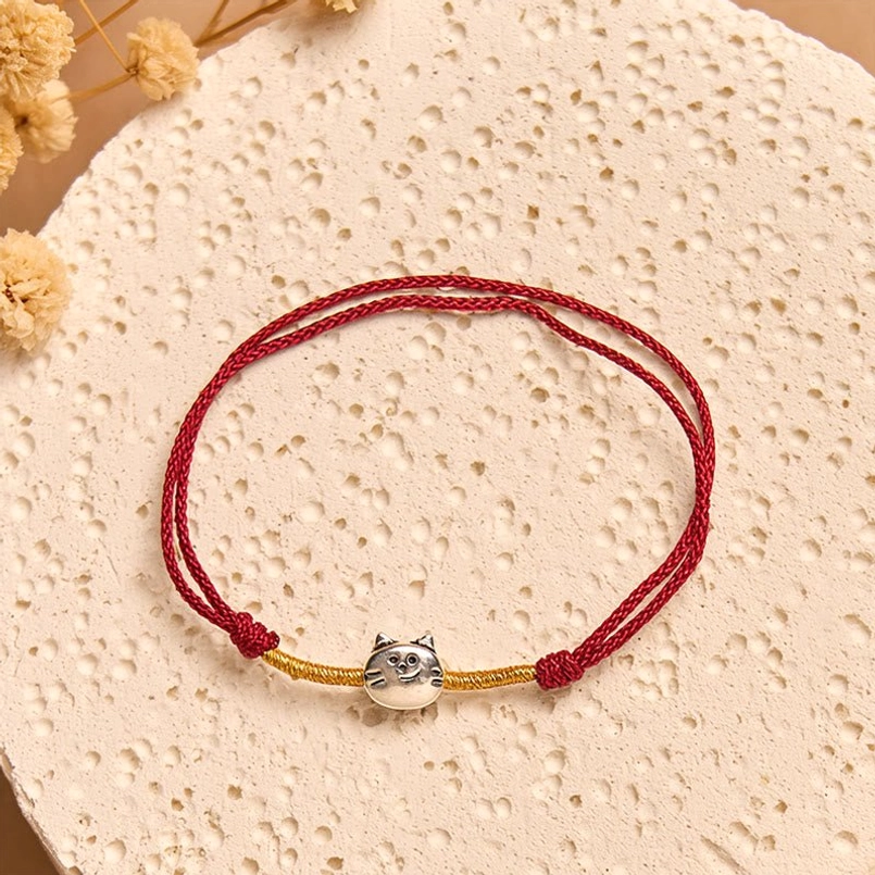 Red string bracelet with silver cat head charm and gold accents on decorative floral plate