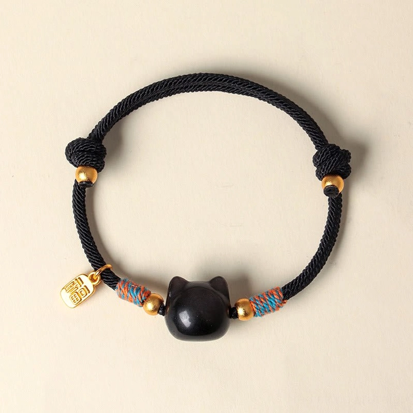 Side angle view of black braided cord cat bracelet with obsidian cat bead and Fu character pendant on beige tabletop