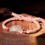 Chalcedony cat bracelet with pink braided string on wooden surface, showing adjustable knot and stone pendant