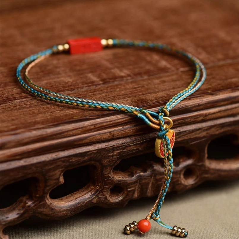 Close-up top-down view of cinnabar blessing bracelet showing teal woven rope, rectangular cinnabar pendant, gold metal beads, and adjustable tassel