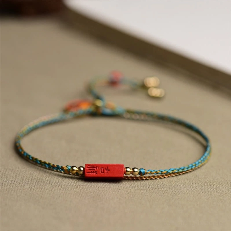 Front close-up of handmade cinnabar blessing bracelet showing blue-yellow woven rope, ji-xiang plaque, and gold metal beads