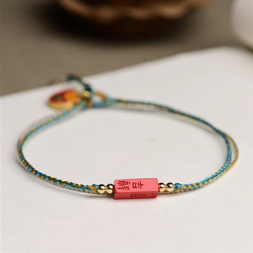 Three-quarter side view of handmade cinnabar blessing bracelet with ji-xiang plaque and enamel charm on book page with dried flowers