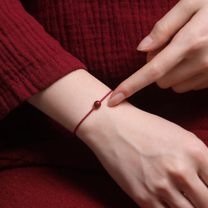Adjusting cinnabar blessing bracelet on wrist with hands in maroon clothing