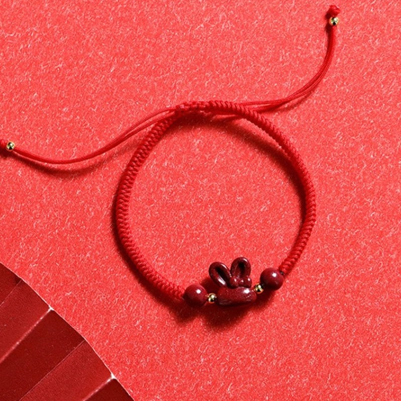 Top-down flat lay of handmade cinnabar rabbit red string bracelet on bright red fabric, full product view with details
