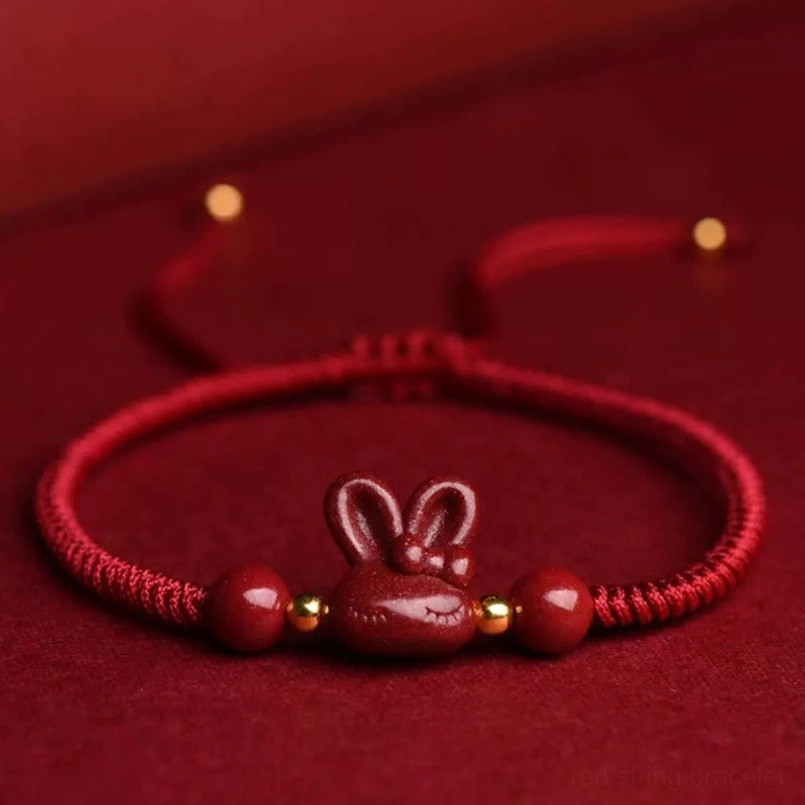 Handmade cinnabar rabbit red string bracelet flat lay on dark red fabric background, showing braided rope cord and gold spacer beads