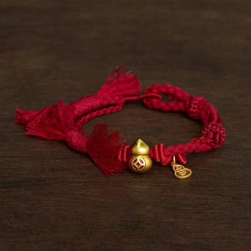 Dark red braided red string bracelet with gold gourd coin charm on dark wooden table