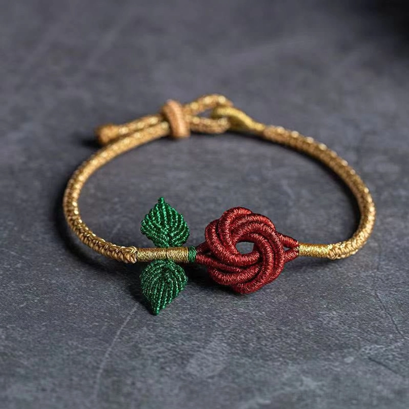 Gold-toned eight thread peace knot red rose bracelet with green leaves on grey stone surface