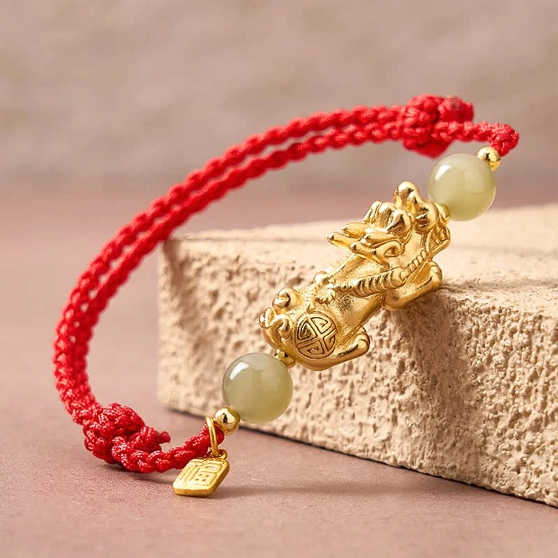 Three quarter view of detailed gold Pi Xiu red string bracelet on beige stone block