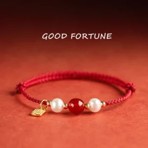 Fu Character red string bracelet with gold charm, red agate bead and white pearls on reddish-brown fabric background