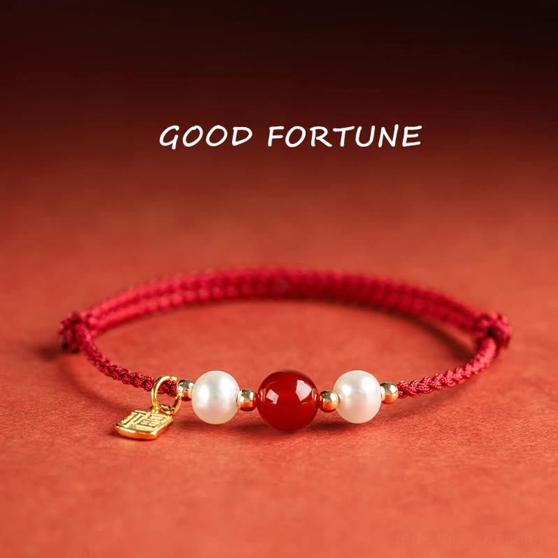 Fu Character red string bracelet with gold charm, red agate bead and white pearls on reddish-brown fabric background