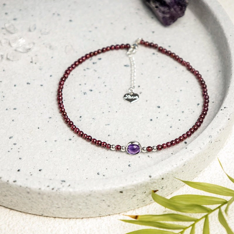 Red string anklet with purple gem on ceramic tray with natural leaf decor