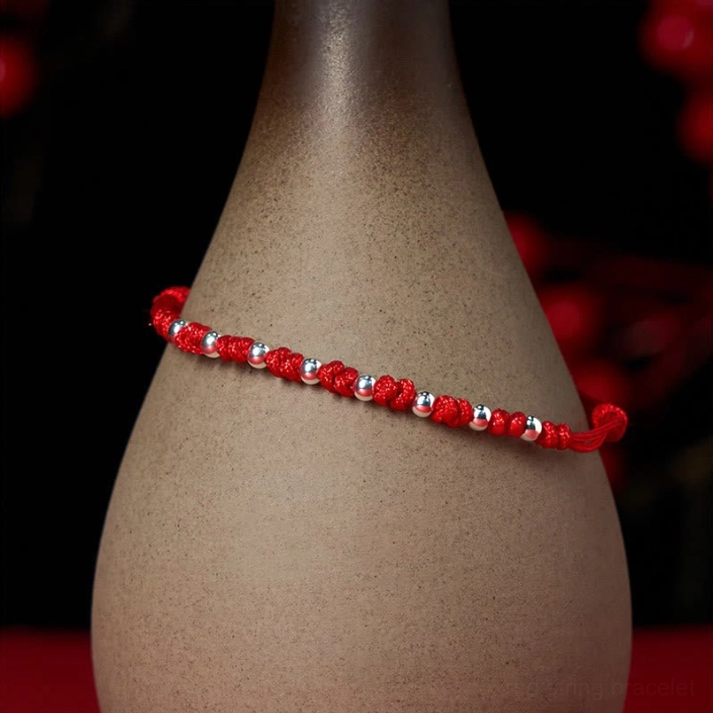 Red cord woven bracelet with silver metal beads arranged along rope with ceramic vase backdrop