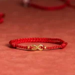 High side perspective of braided red string bracelet with brass infinity charm on red matte tabletop