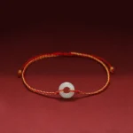 Jade peace buckle red string braided bracelet with gold-toned cord and white jade donut pendant on dark red background