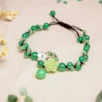Green jade prosperity anklet with braided cord and carved rose flower charm, displayed on beige surface with dried flowers