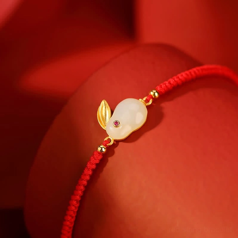 Hetian white jade rabbit charm bracelet with gold ear accents and red cord on solid background