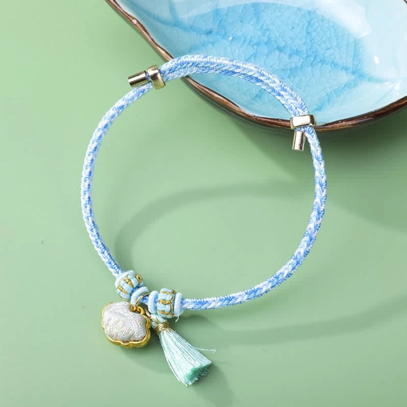 Light blue five-color braided cord luck bracelet with shell charm and green Song brocade tassel flat lay