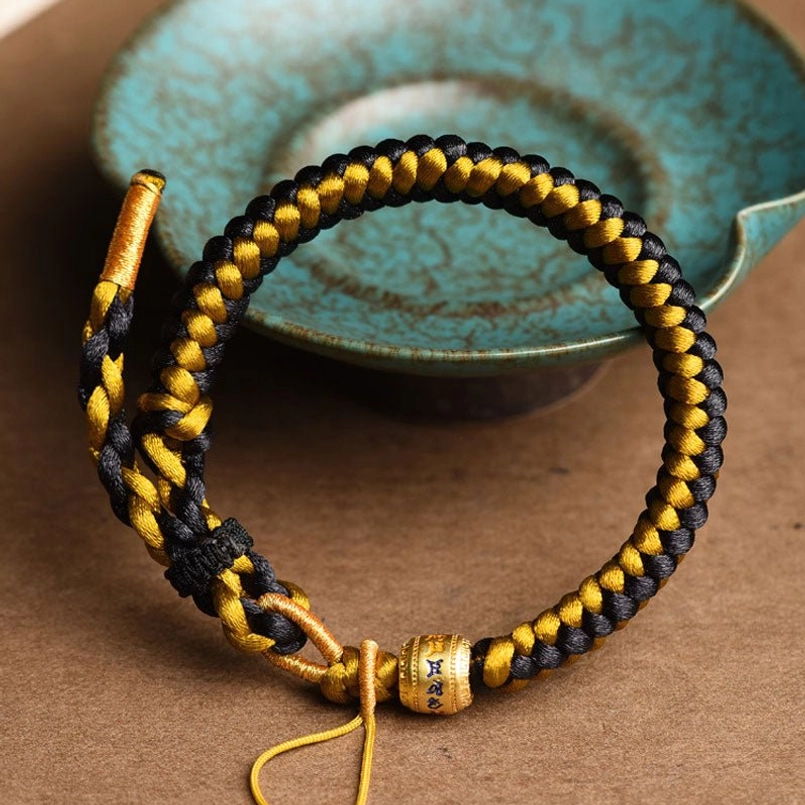 Sacred mantra bracelet angled top-down shot displaying black-gold braided cord and metal charm accent