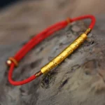 Tibetan Om Mani Padme Hum red string bracelet with gold-plated sterling silver engraved tube bead on wooden surface