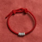 Om Mani Padme Hum red string protection bracelet with silver mantra bead, top-down view on dark fabric background