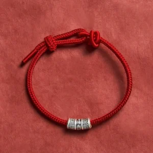 Om Mani Padme Hum red string protection bracelet with silver mantra bead, top-down view on dark fabric background