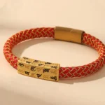 Om Mani Padme Hum Tibetan blessing bracelet with red woven rope, gold thread, and engraved Sanskrit charm on beige studio background