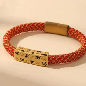Om Mani Padme Hum Tibetan blessing bracelet with red woven rope, gold thread, and engraved Sanskrit charm on beige studio background