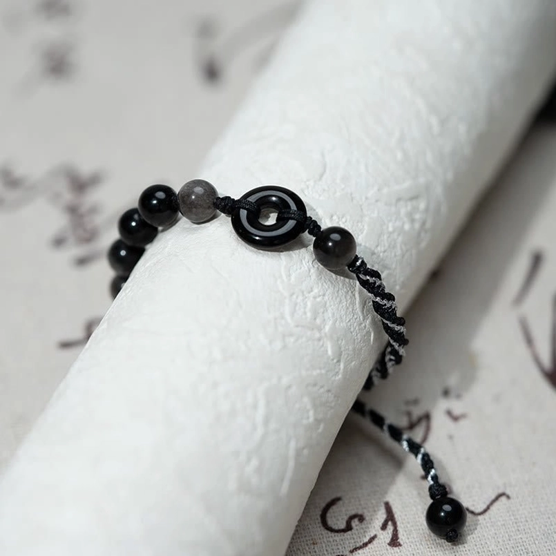 Close-up side view of black obsidian peace buckle bracelet featuring black and white woven rope on traditional scroll background