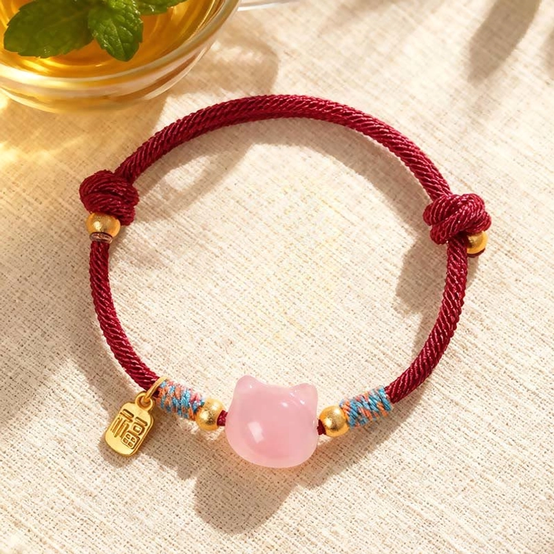 Pink crystal cat protection bracelet with red rope and Fu character charm displayed on linen tabletop with tea cup