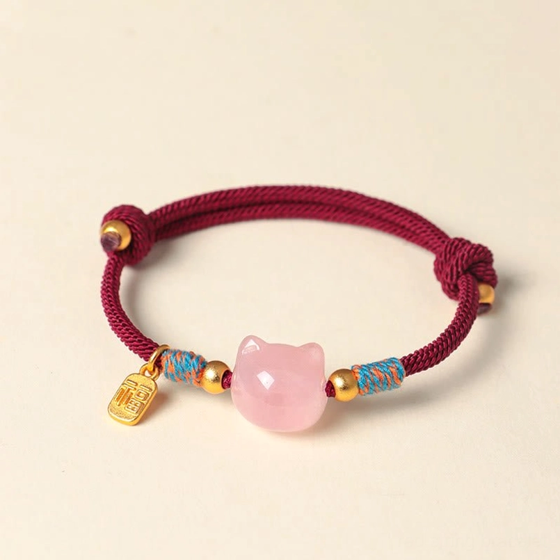 Overhead view of burgundy rope cat bracelet featuring pink crystal bead and Fu symbol charm on light pink background