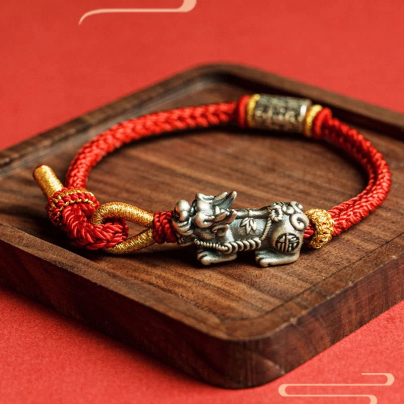 Handwoven red and gold rope Pixiu bracelet with sterling silver charm on wooden coaster, flat lay top view