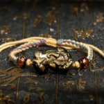 Gold Sheen Obsidian PiXiu wealth bracelet displayed overhead with braided rope on dark background with Chinese patterns