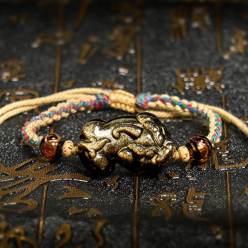 Gold Sheen Obsidian PiXiu wealth bracelet displayed overhead with braided rope on dark background with Chinese patterns