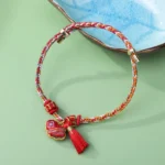 Red five-color braided cord luck bracelet with embroidered heart charm and red Song brocade tassel on ceramic dish