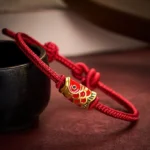Red koi fish braided string bracelet with gold-red enamel charm, adjustable cord knot, close-up three-quarter view