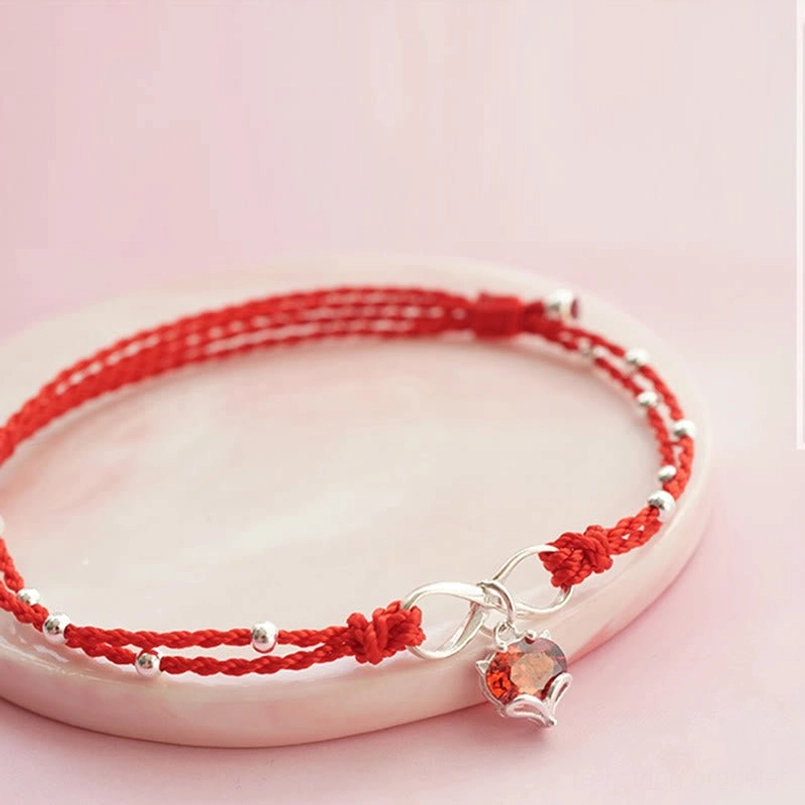 Red string infinity ankle bracelet with silver clasp and heart pendant, side flat lay on pink ceramic tray