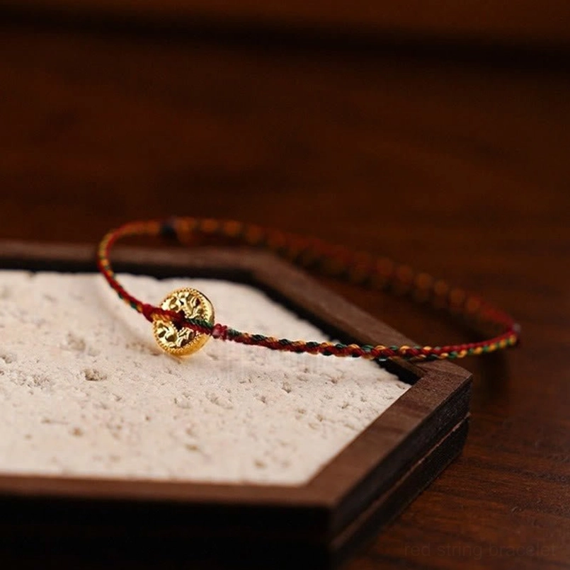 Handmade woven red string anklet with gold copper coin charm displayed on dark wood tray