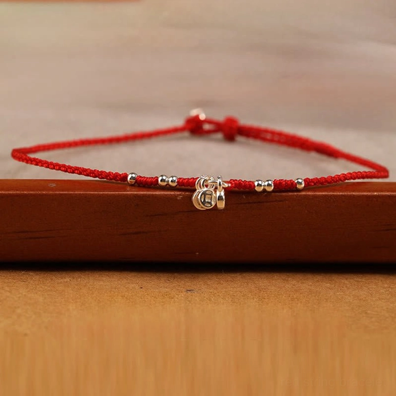 Red string ankle bracelet on wooden surface featuring double silver copper coin charms and beads