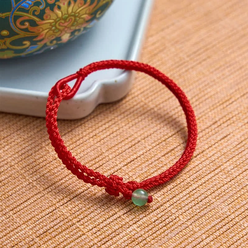 Red string bracelet with green aventurine bead and adjustable clasp on woven table mat and ceramic tray