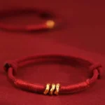 999 gold bead red string bracelet with three twisted gold rings on burgundy background