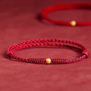 Dark red braided string bracelet with gold accent bead, close-up on fabric