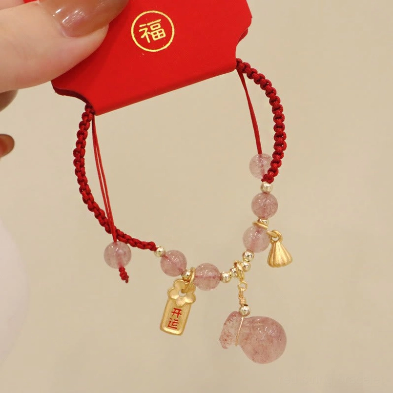 Braided strawberry quartz red string bracelet held in hand with lucky fortune envelope background showing gold-toned lotus pod charm and money bag quartz pendant