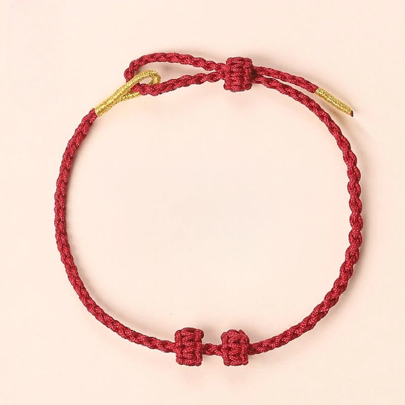 Minimalist red braided bracelet with decorative knots and gold cord ends on pink background