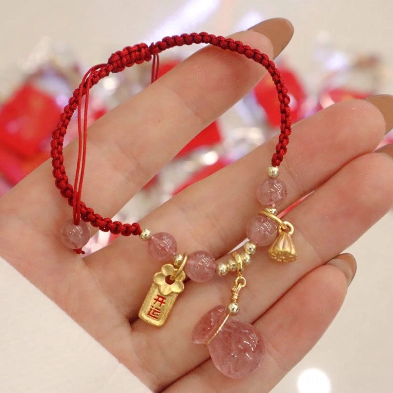 Strawberry quartz red string bracelet close up in palm with festive red blurred background showing pink quartz stone, braided red rope, gold lotus pod charm and lucky fortune tag