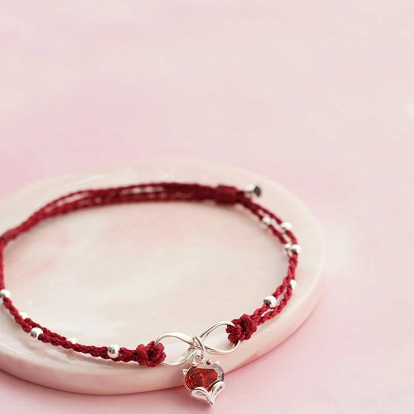 Double-layered red braided anklet on pink tray showing braid texture, silver beads, and heart pendant detail