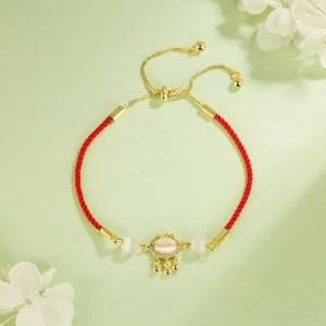 Red string cat's eye lock charm bracelet with gold-plated hardware on light green background with white flowers, adjustable pull-tie clasp visible